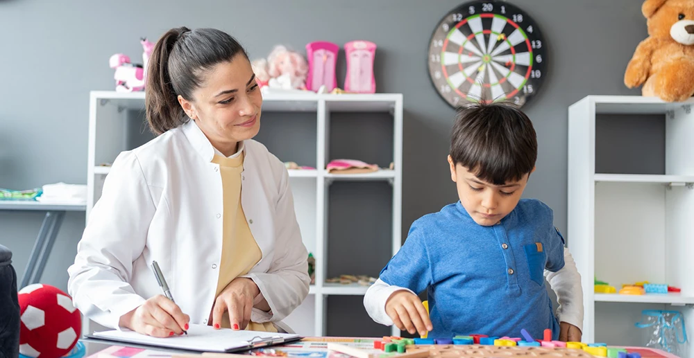Woman with a clipboard watching a child work on a puzzle.